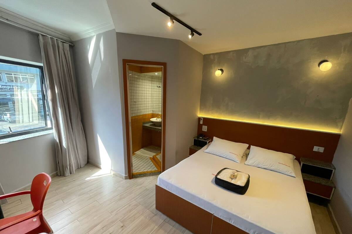 Une chambre d’hôtel comprenant un lit, des tables de chevet, une chaise, une fenêtre avec des rideaux et une salle de bain visible à travers une porte en verre.
