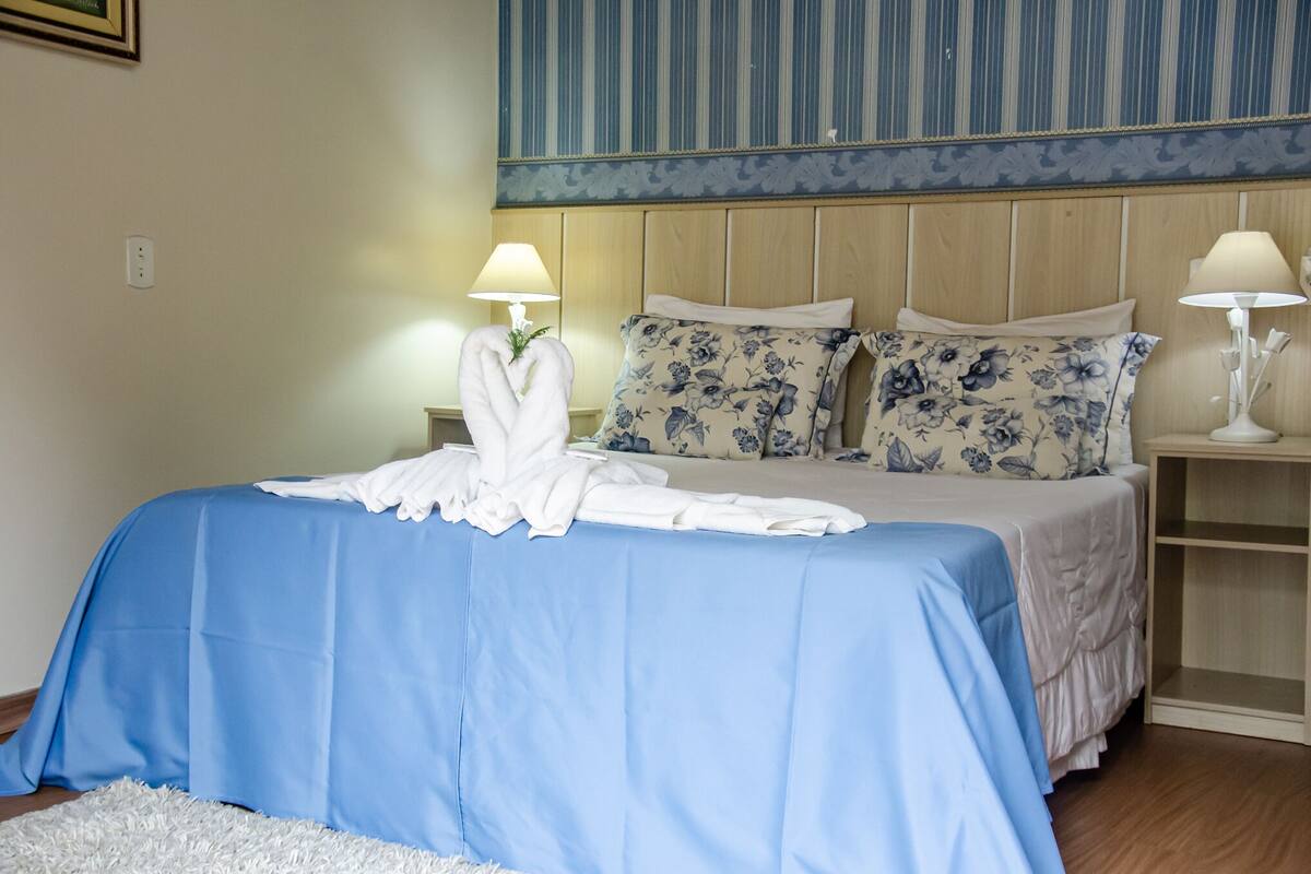 A neatly made bed with a blue bedspread, two floral pillows, and a white towel on the side. There are two bedside lamps and a small wooden nightstand with a shelf.