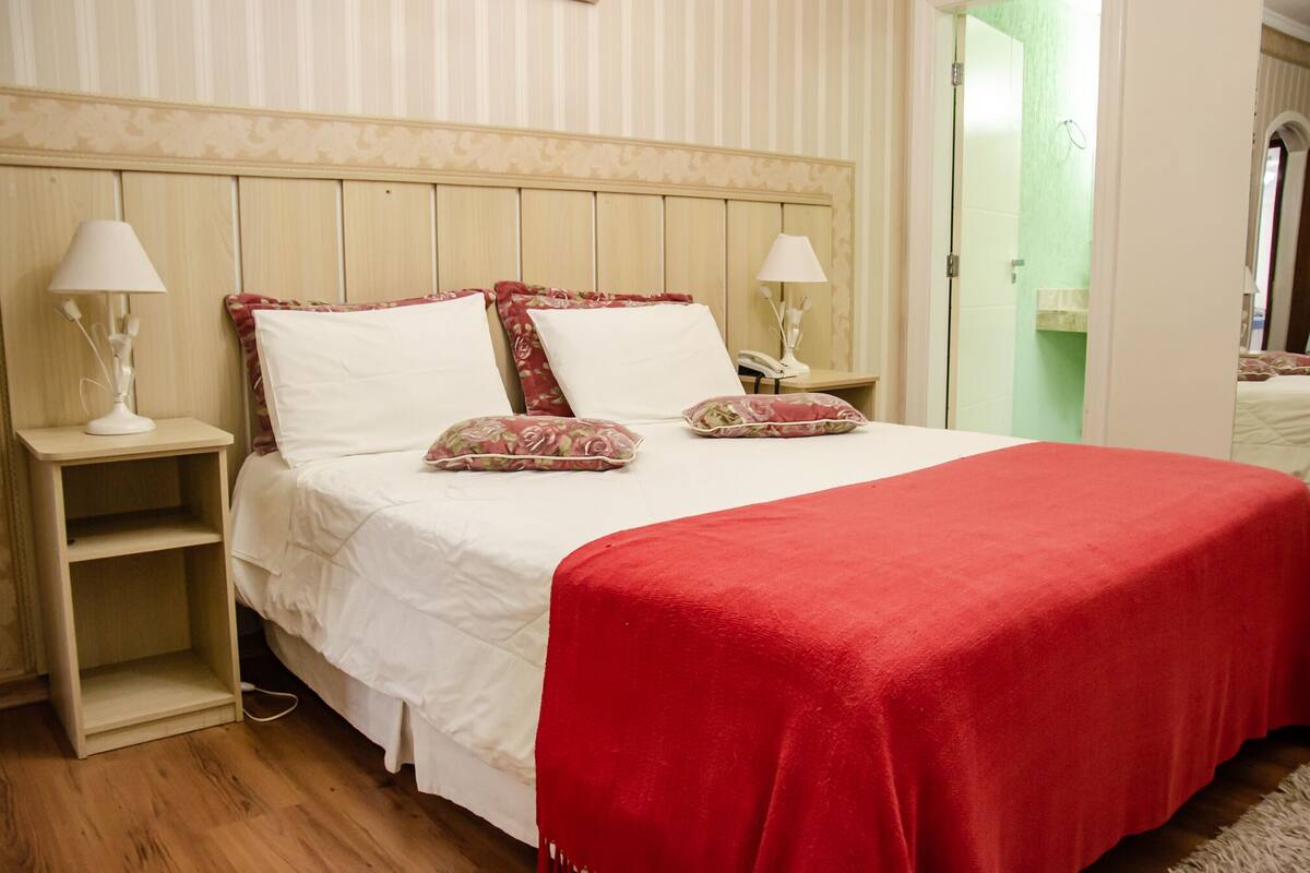 A neatly made bed with a red blanket, white pillows, and floral-patterned pillows. There are two bedside tables with lamps and a telephone. The room has wooden paneling on the walls and a green door in the background.