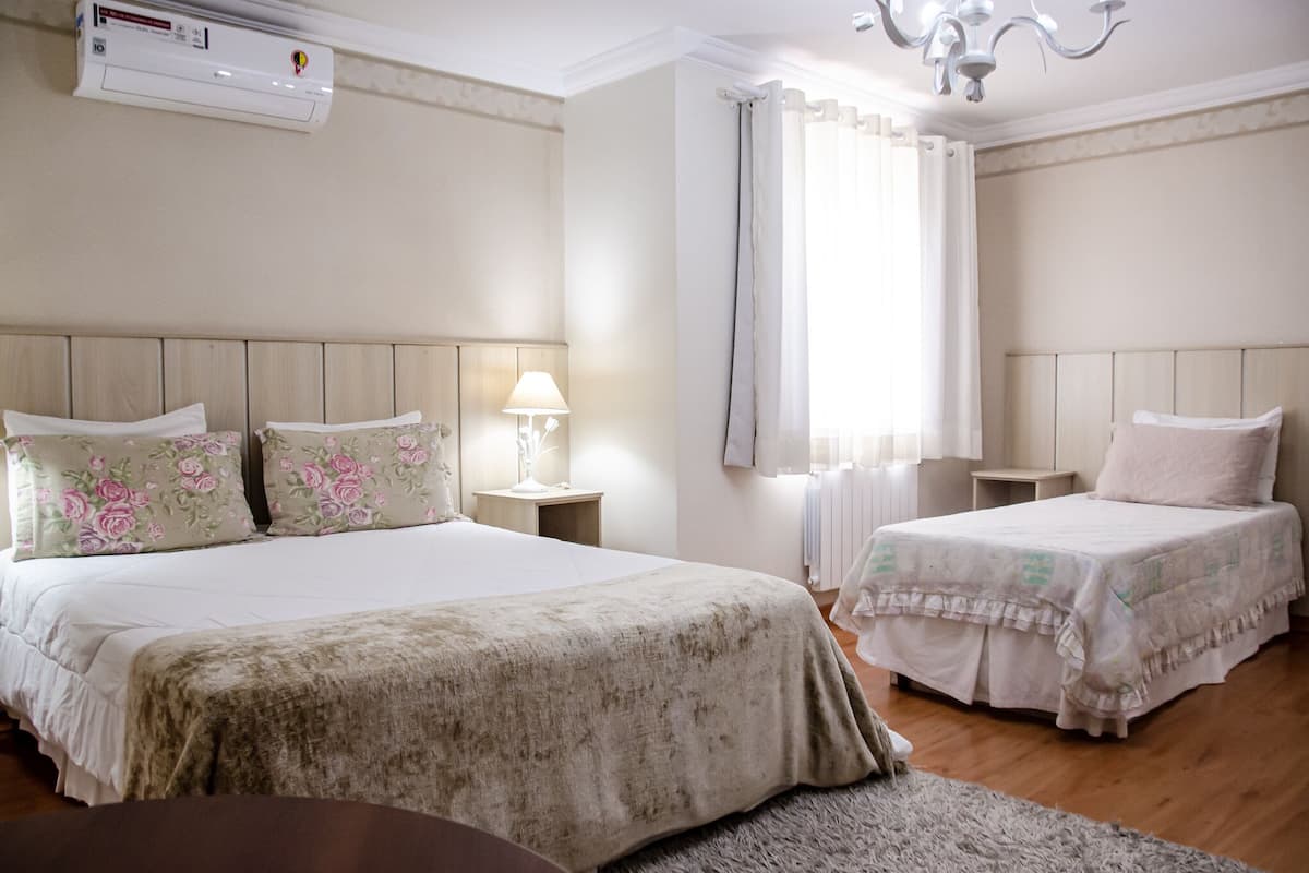 A hotel room with two beds, a chandelier, an air conditioner, and a window with curtains.