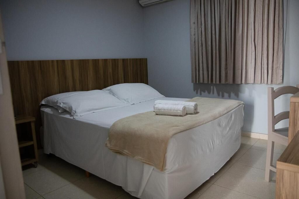 Una cama individual con ropa blanca, un edredón color beige y una toalla enrollada sobre la cama.