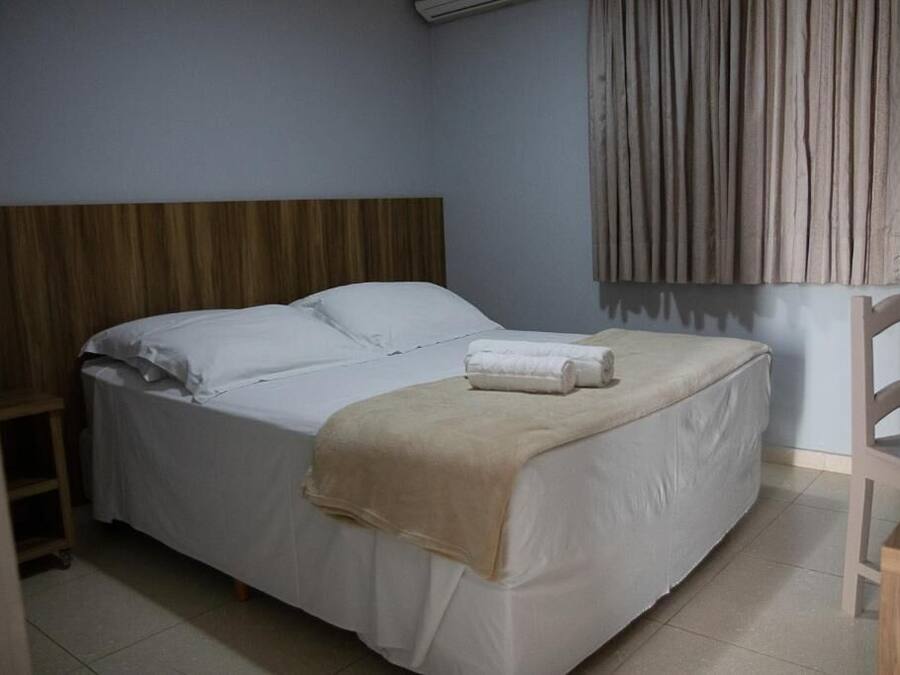 Una cama individual con ropa blanca, un edredón color beige y una toalla enrollada sobre la cama.