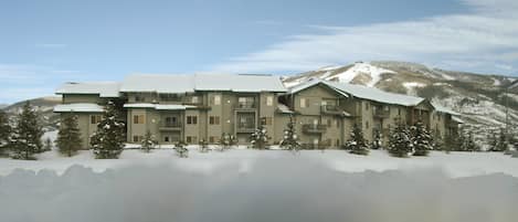 Un grand hôtel à plusieurs étages, dont le toit est recouvert de neige et qui possède de nombreux balcons, entouré d’arbres et de montagnes également couverts de neige.