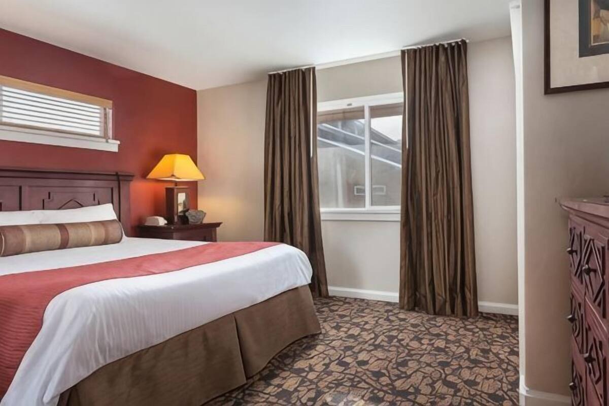 A hotel room with a bed, a nightstand with a lamp, a dresser, a window with curtains, and a patterned carpet.