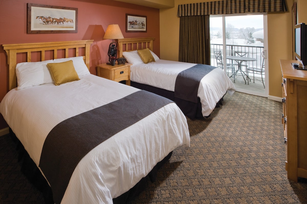 A hotel room with two beds, a wooden headboard, a nightstand with a lamp, a television, and a balcony with outdoor seating.