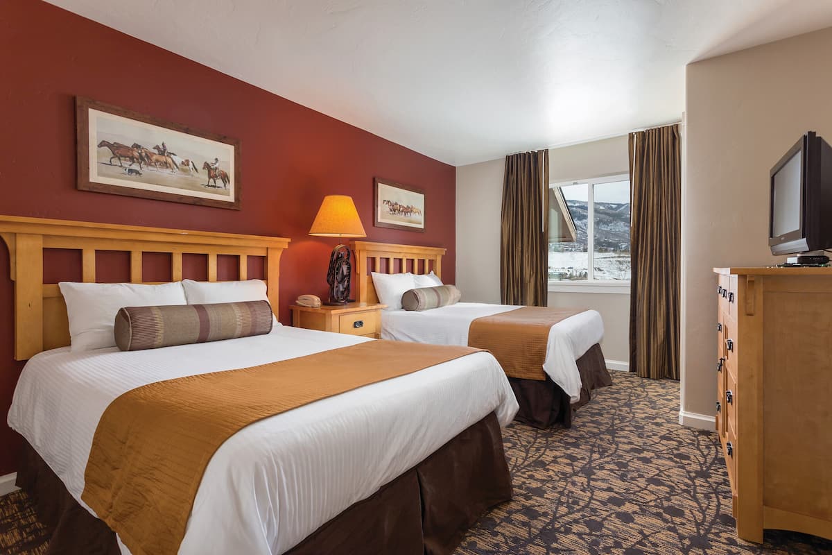 A hotel room with two beds, a wooden headboard, a lamp, a dresser, a television, and a window with curtains.