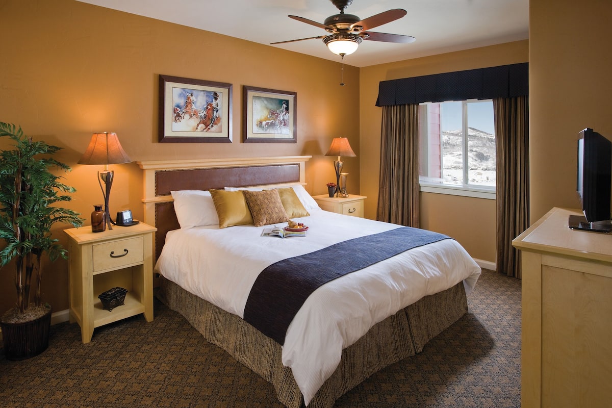 A hotel room with a large bed, two bedside lamps, a ceiling fan, a TV, and a window with curtains.