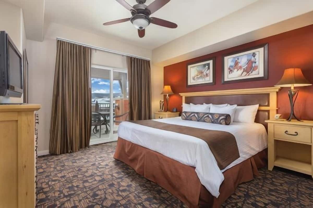 A hotel room with a bed, a ceiling fan, a TV, a desk, a chair, a window with curtains, and a painting on the wall.