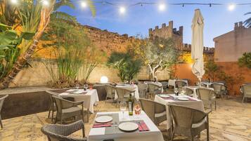 An outdoor dining area with tables and chairs, string lights, and a backdrop of a stone wall and greenery.