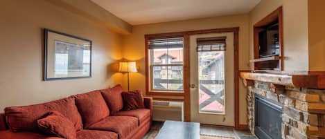 Ein gemütliches Wohnzimmer mit einem roten Sofa, einem Couchtisch, einem Kamin und einem großen Fenster mit Blick auf Gebäude.