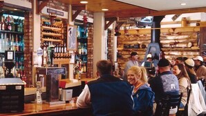 A bar area with wooden shelves stocked with bottles, a cash register, and a television screen. - Highland House (Snowshoe)