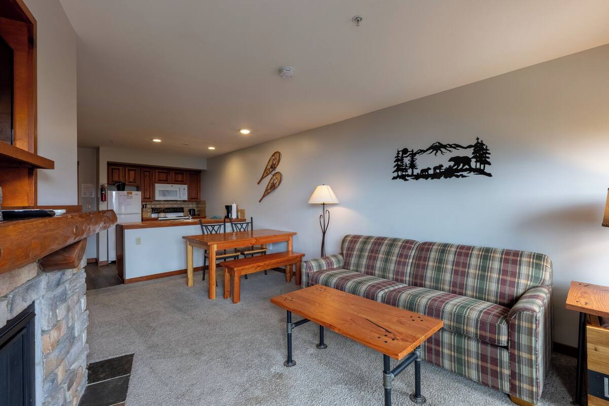 A cozy living room with a fireplace, a plaid sofa, a wooden coffee table, and a dining area with a wooden table and benches.