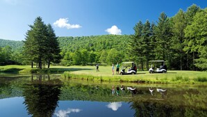 A golf course with a pond reflecting trees and people, surrounded by a forested area. - Highland House (Snowshoe)