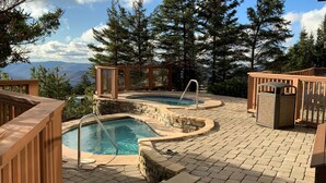 A pool area with two hot tubs, wooden deck, stone steps, and a view of mountains.