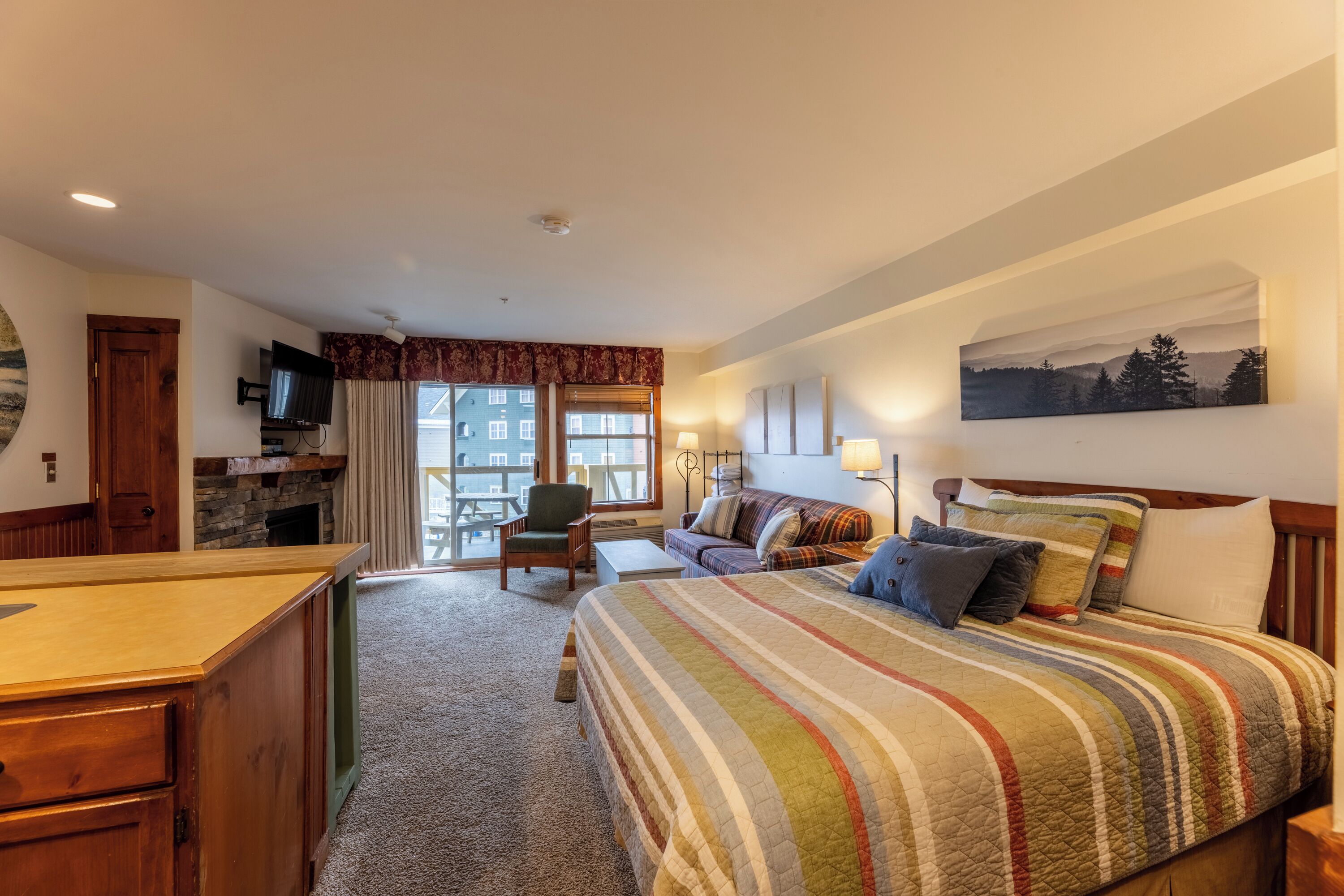 A hotel room with a bed, a desk, a chair, a TV, a fireplace, and a large window.