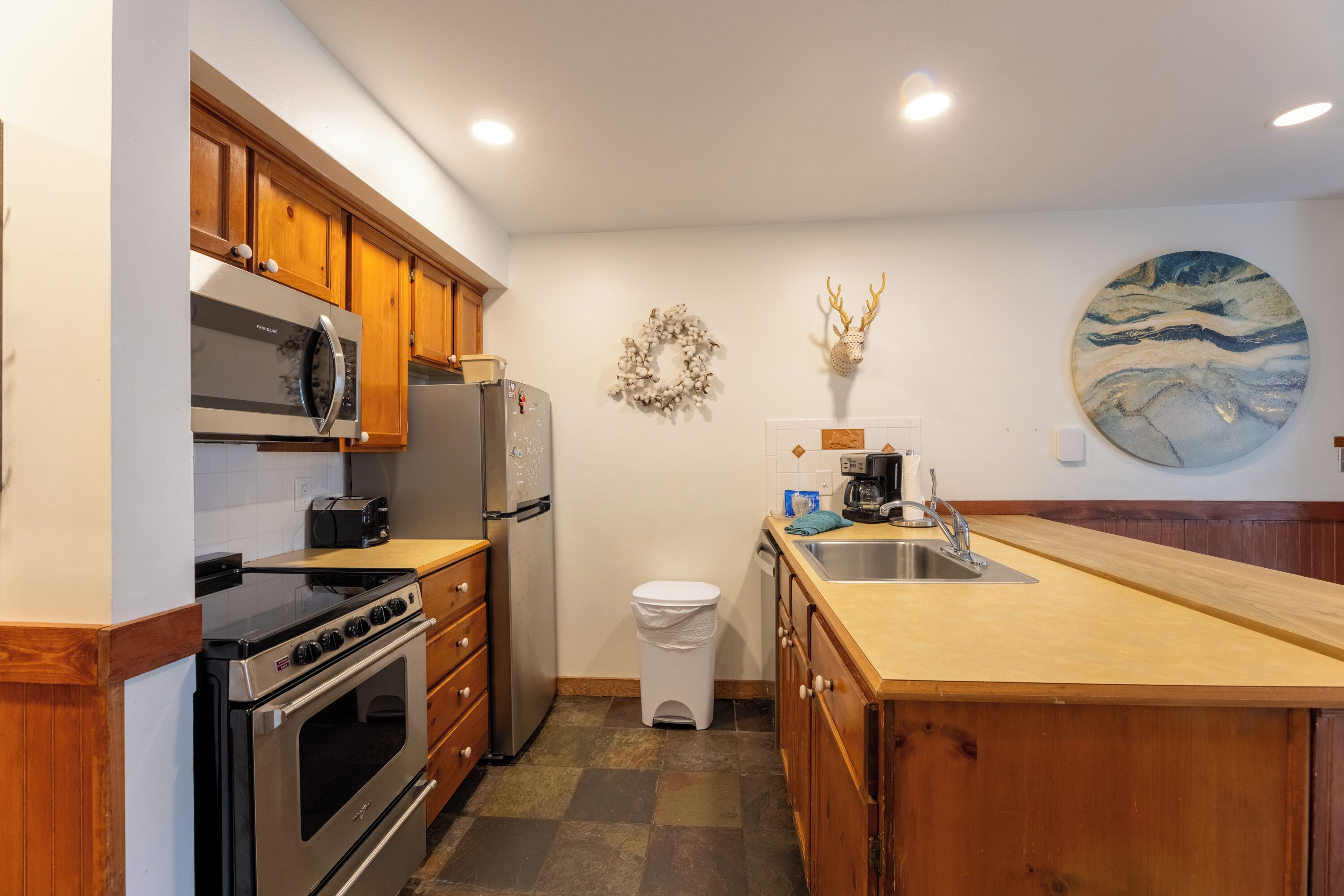 A kitchen with wooden cabinets, stainless steel appliances, a white trash bin, a sink, and a decorative wreath and wall art.