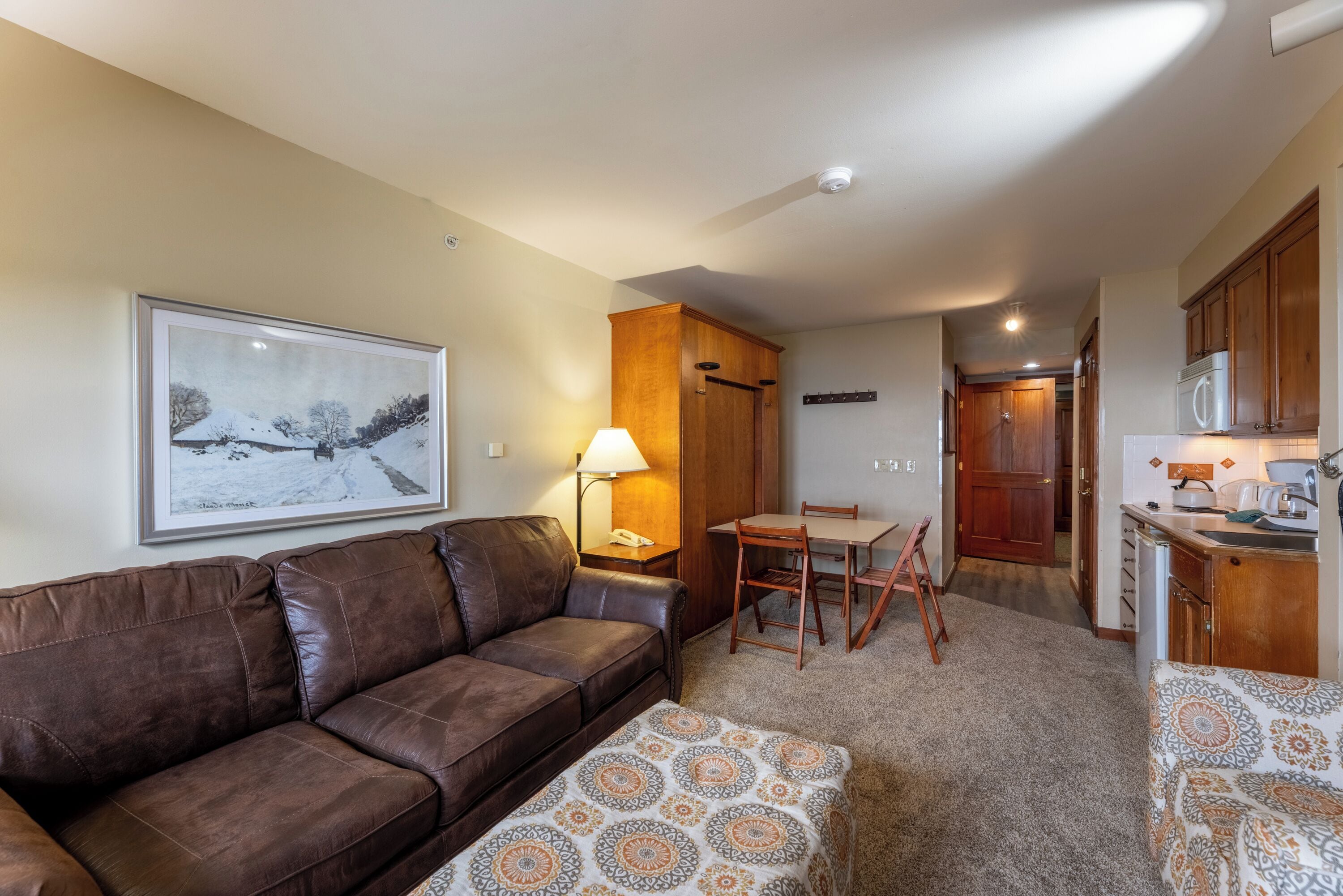 A cozy living room with a brown leather sofa, a patterned ottoman, a dining table with chairs, and a kitchen area with wooden cabinets and a microwave.