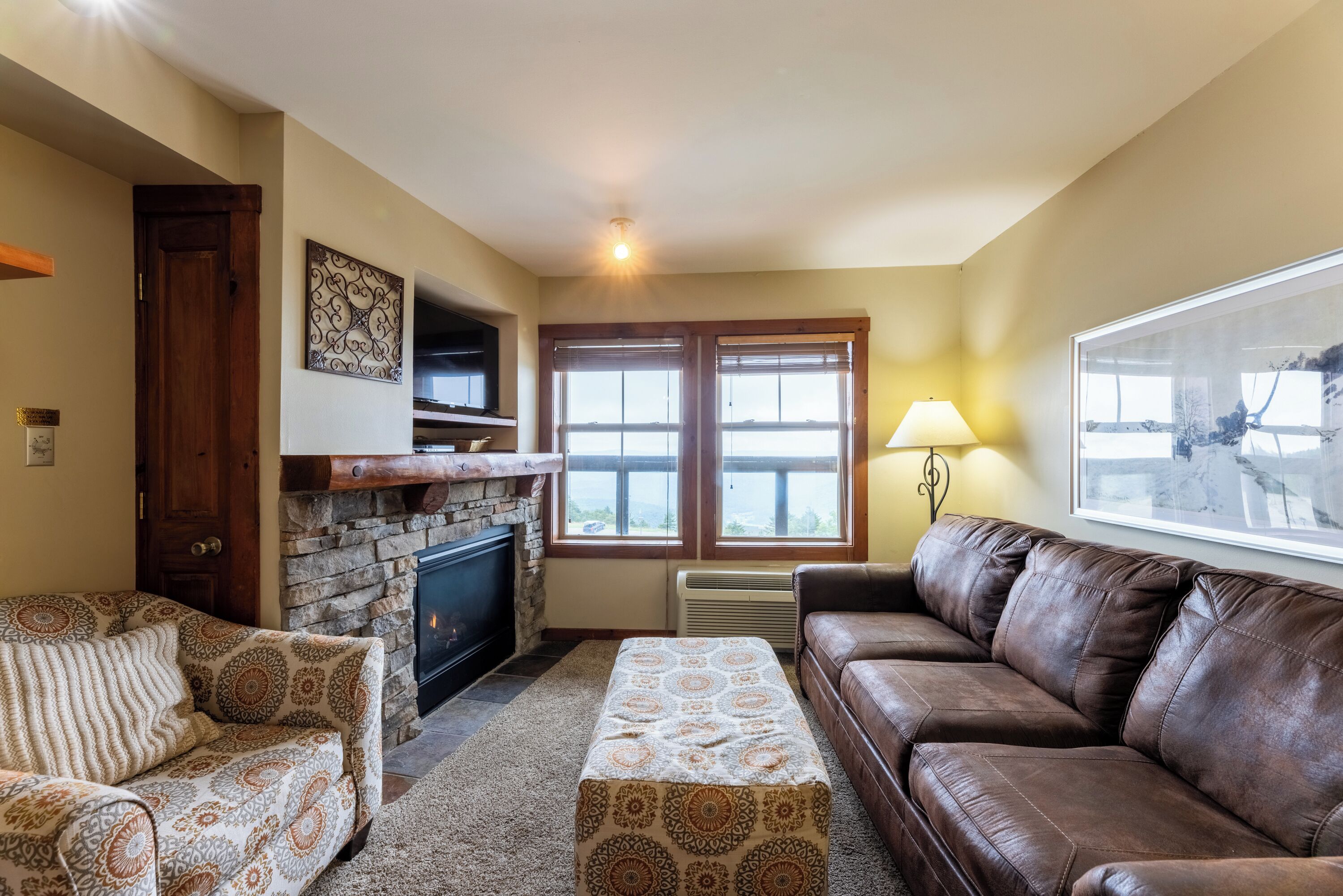 A living room with a fireplace, a sofa, a patterned ottoman, and a lamp.