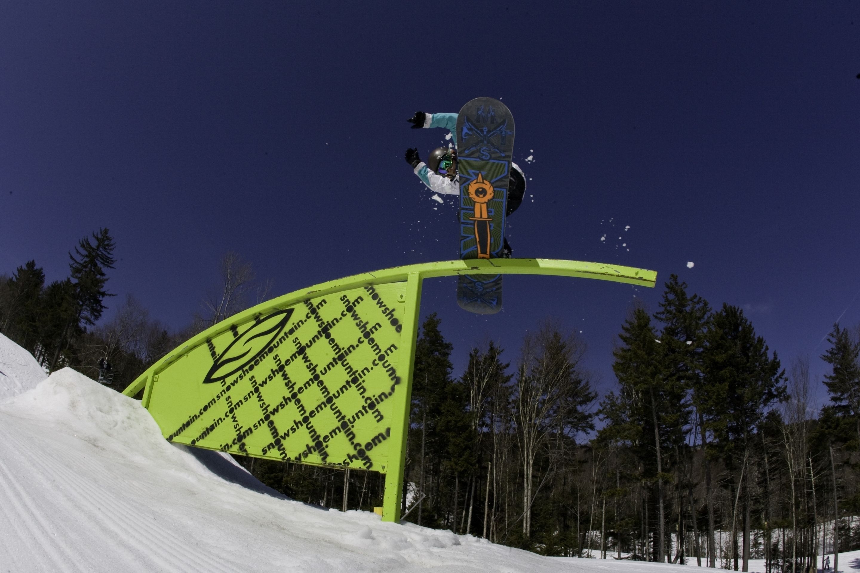 A snowboarder performing a jump over a green structure with a logo and text.
