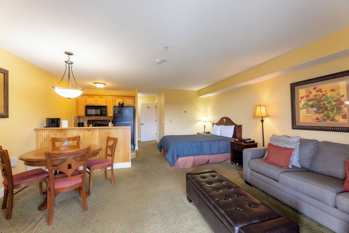 A compact living space with a kitchenette, a dining area, a bedroom with a bed and nightstand, and a seating area with a sofa and ottoman.