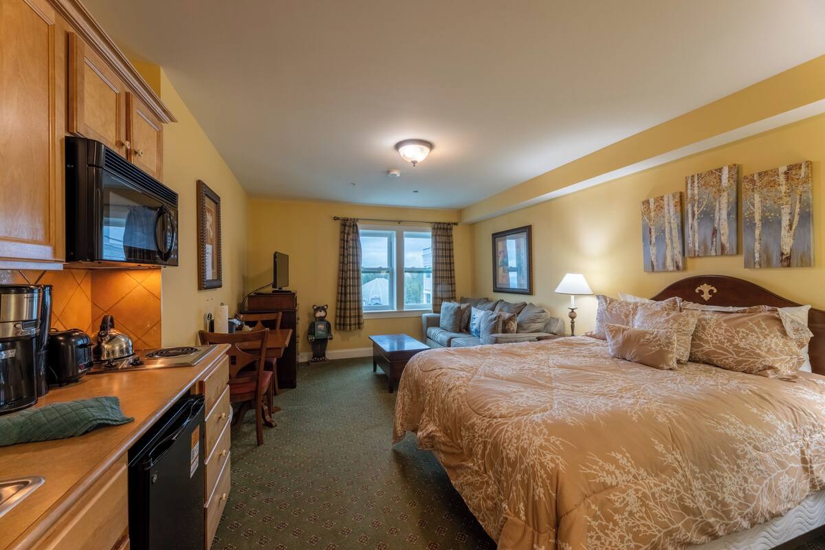 A hotel room with a kitchenette, a bed with a floral bedspread, a sofa, a window with curtains, and artwork on the walls.