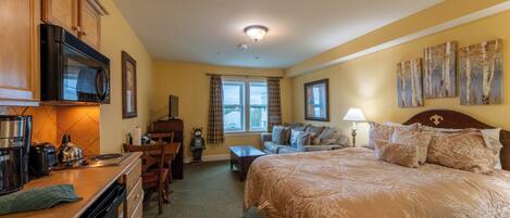 A hotel room with a kitchenette, a bed with a floral bedspread, a sofa, a window with curtains, and artwork on the walls.