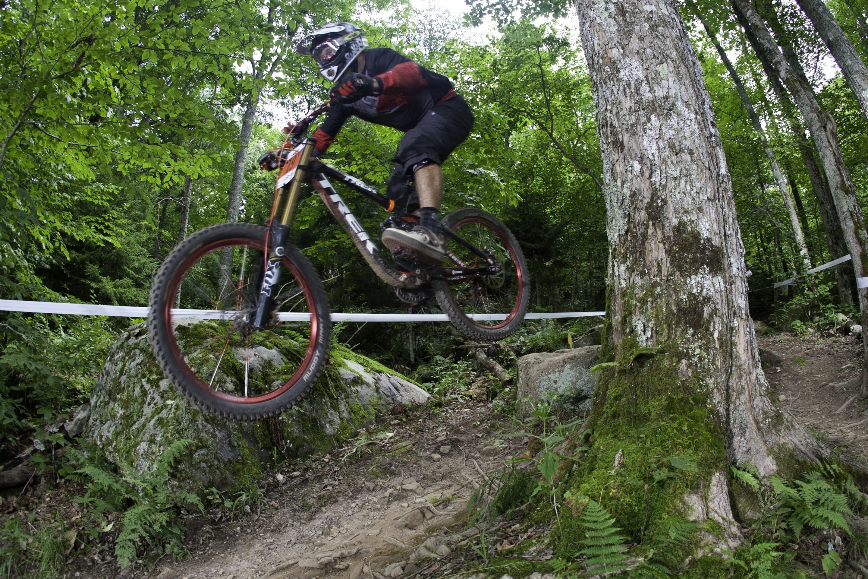 A mountain biker navigating a dirt trail in a forested area with trees and natural terrain.