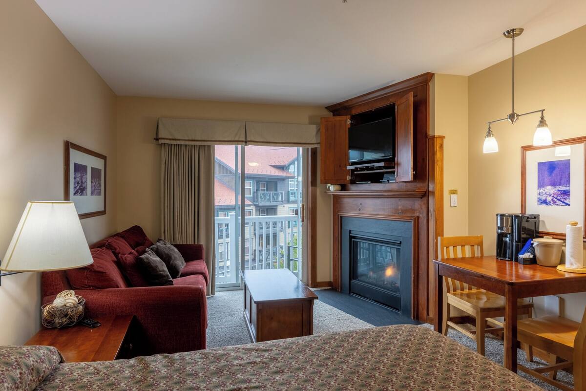 A hotel room with a sofa, a small table, a fireplace, a television, and a balcony with a view of buildings.