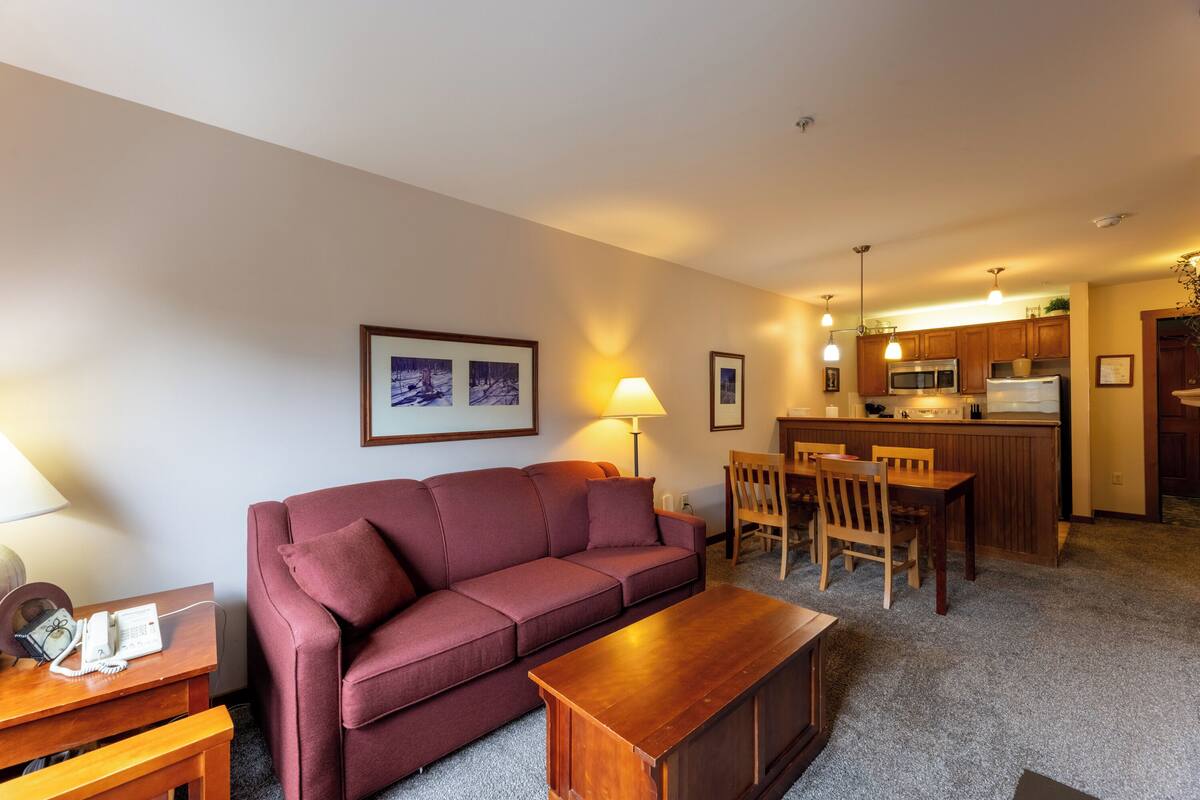 A living room with a sofa, a wooden coffee table, and a dining area with chairs and a table. There is a kitchen area with a microwave, refrigerator, and a bar counter.