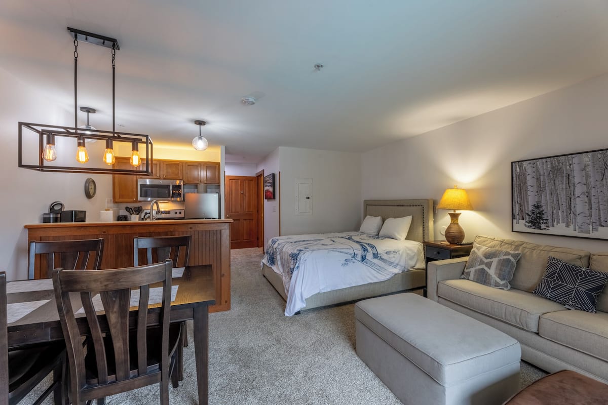 A compact living space with a kitchenette, a dining area, a bedroom with a bed and nightstand, and a sofa with a lamp.