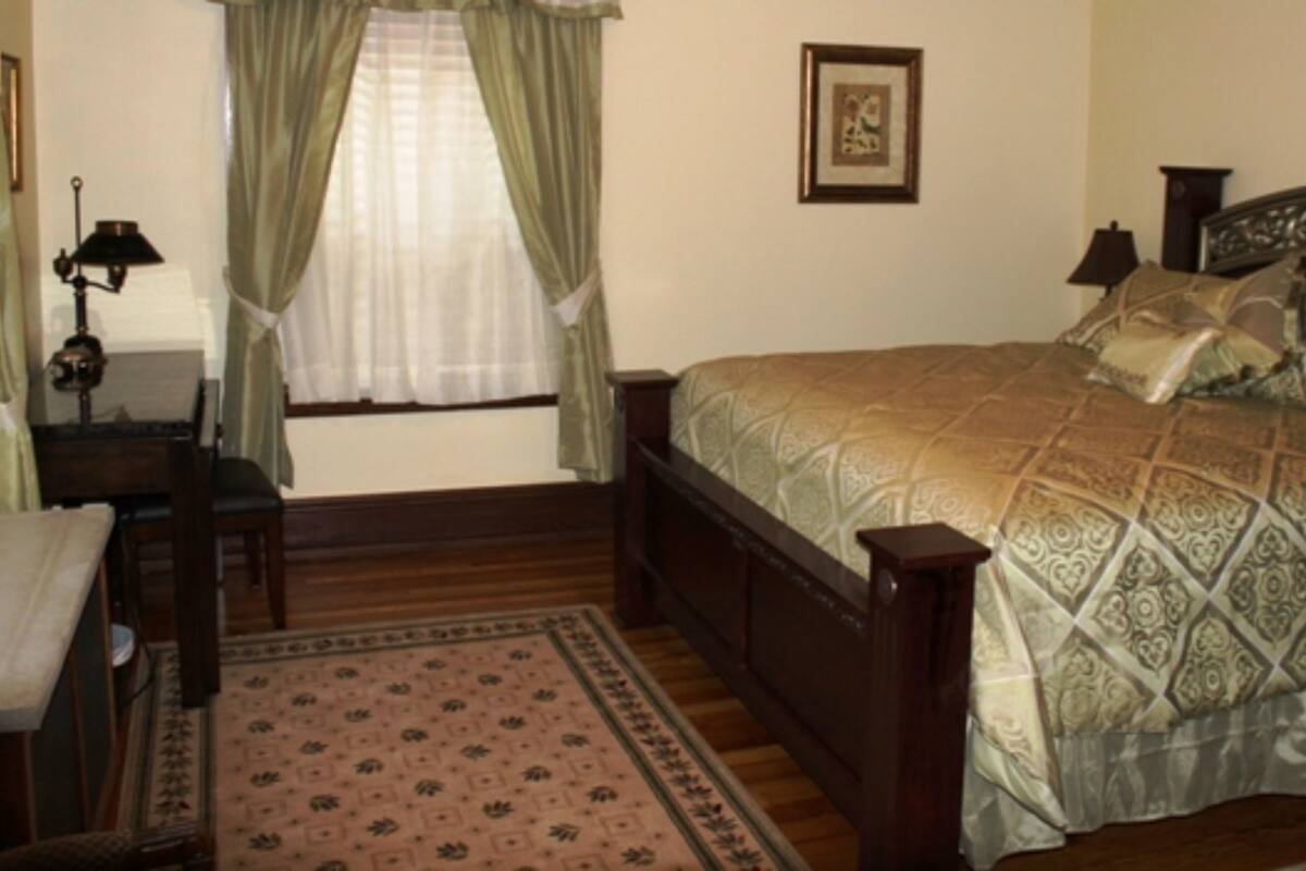 A bedroom with a wooden bed, a nightstand, a lamp, a window with curtains, and a framed picture on the wall.