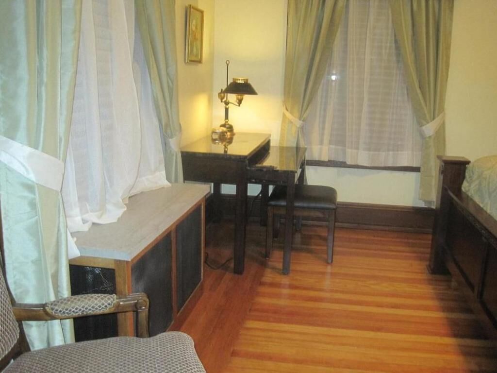 A room with a wooden floor, a desk with a lamp, a chair, and a bed.