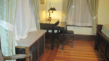 A room with a wooden floor, a desk with a lamp, a chair, and a bed.