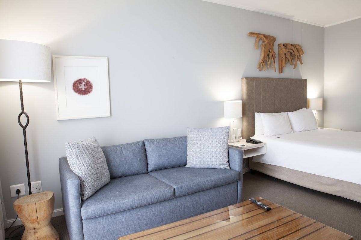 Quarto de hotel com um sofá cinza, uma mesa de centro de madeira, uma cama com roupa de cama branca, uma mesa de cabeceira com luminárias e uma obra de arte pendurada na parede.