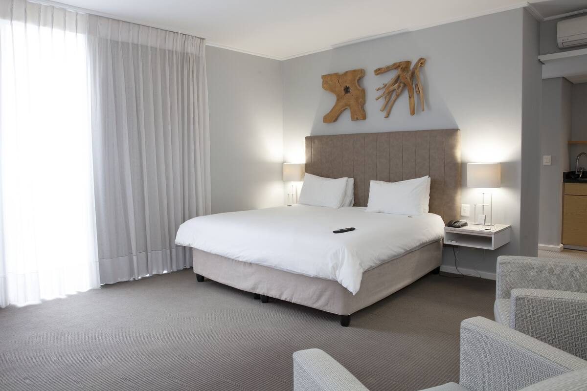 Quarto de hotel com uma cama grande, uma mesa de cabeceira com abajur, cabeceira de madeira e uma janela ampla com cortinas.