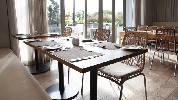 A dining area with tables set for two, wicker chairs, and a view of greenery outside. - Atlantic Beach Hotel (Cape Town)