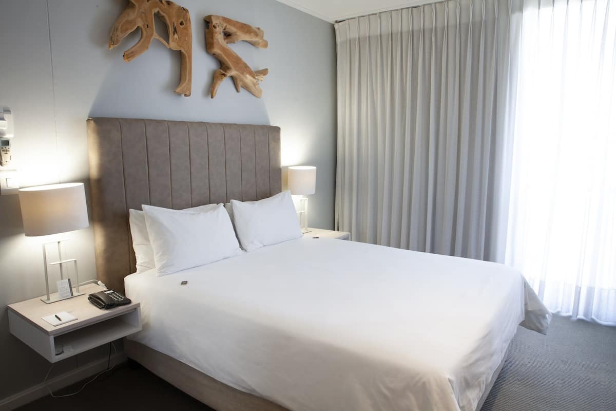 Quarto de hotel com uma cama grande, duas luminárias de cabeceira, um telefone e um objeto decorativo de madeira na parede.