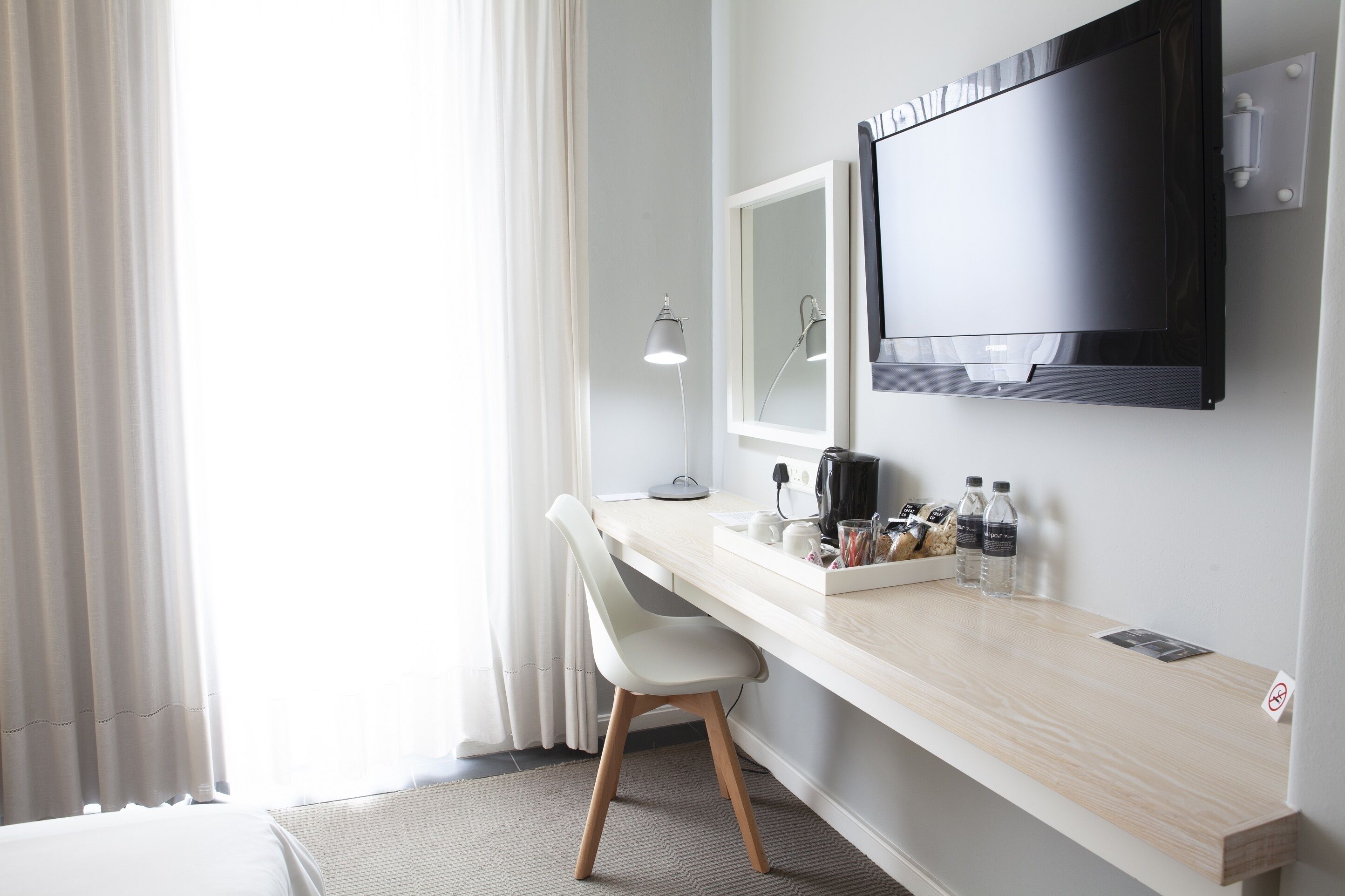 A hotel room with a desk, a chair, a TV, a lamp, and a window with curtains.