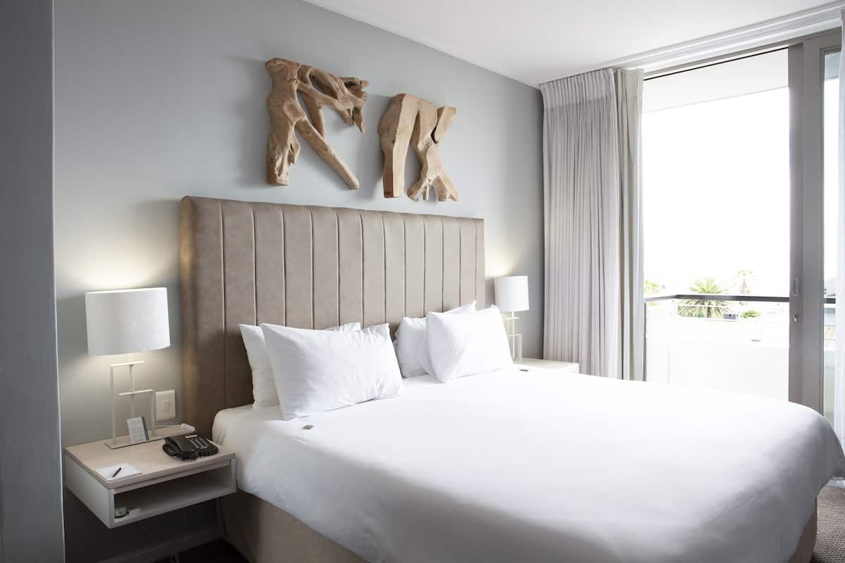 Quarto de hotel com uma cama grande, duas luminárias de cabeceira, mesa de apoio com telefone e um bloco de notas, e uma janela com cortinas.