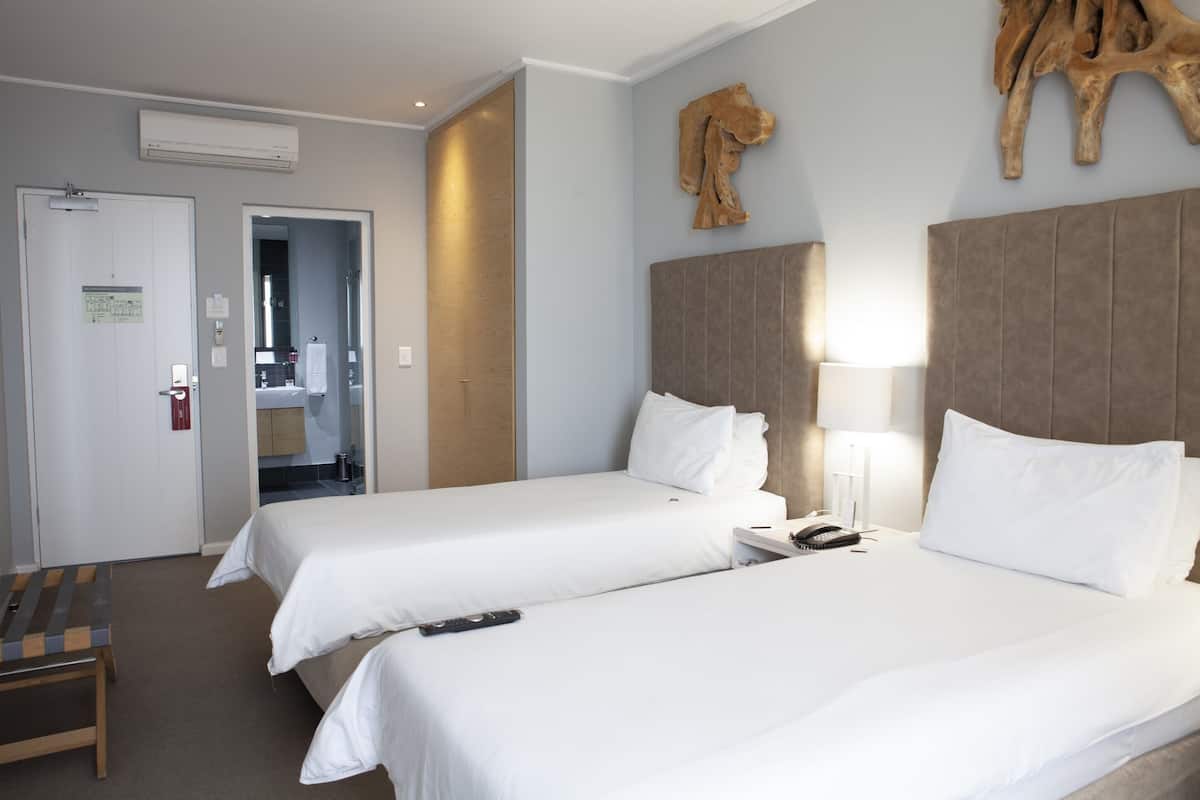 Quarto de hotel com duas camas, um banco de madeira, uma escrivaninha com abajur e uma grande escultura de madeira na parede.