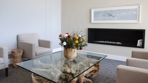 A modern living room with a glass coffee table, beige sofas, a wooden side table, and a fireplace with a framed artwork above it. - Atlantic Beach Hotel (Cape Town)