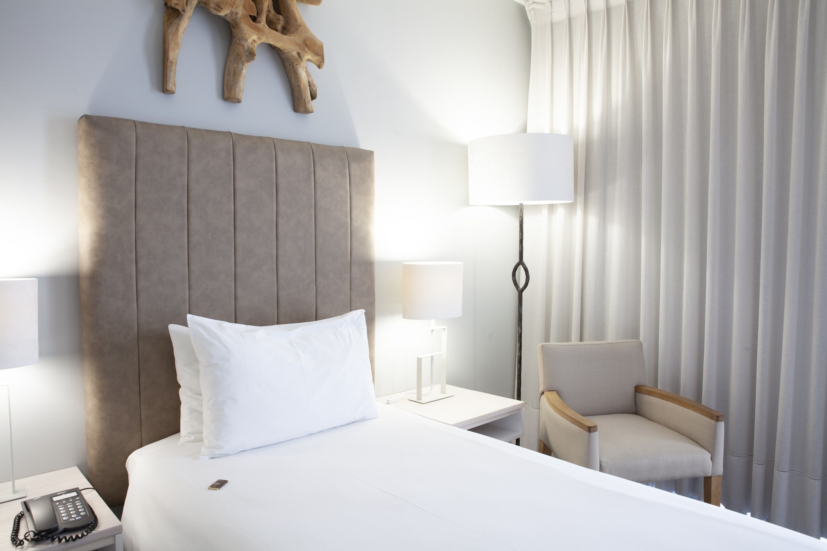 A hotel room with a large bed, a nightstand with a phone, a lamp, a chair, and a curtain.
