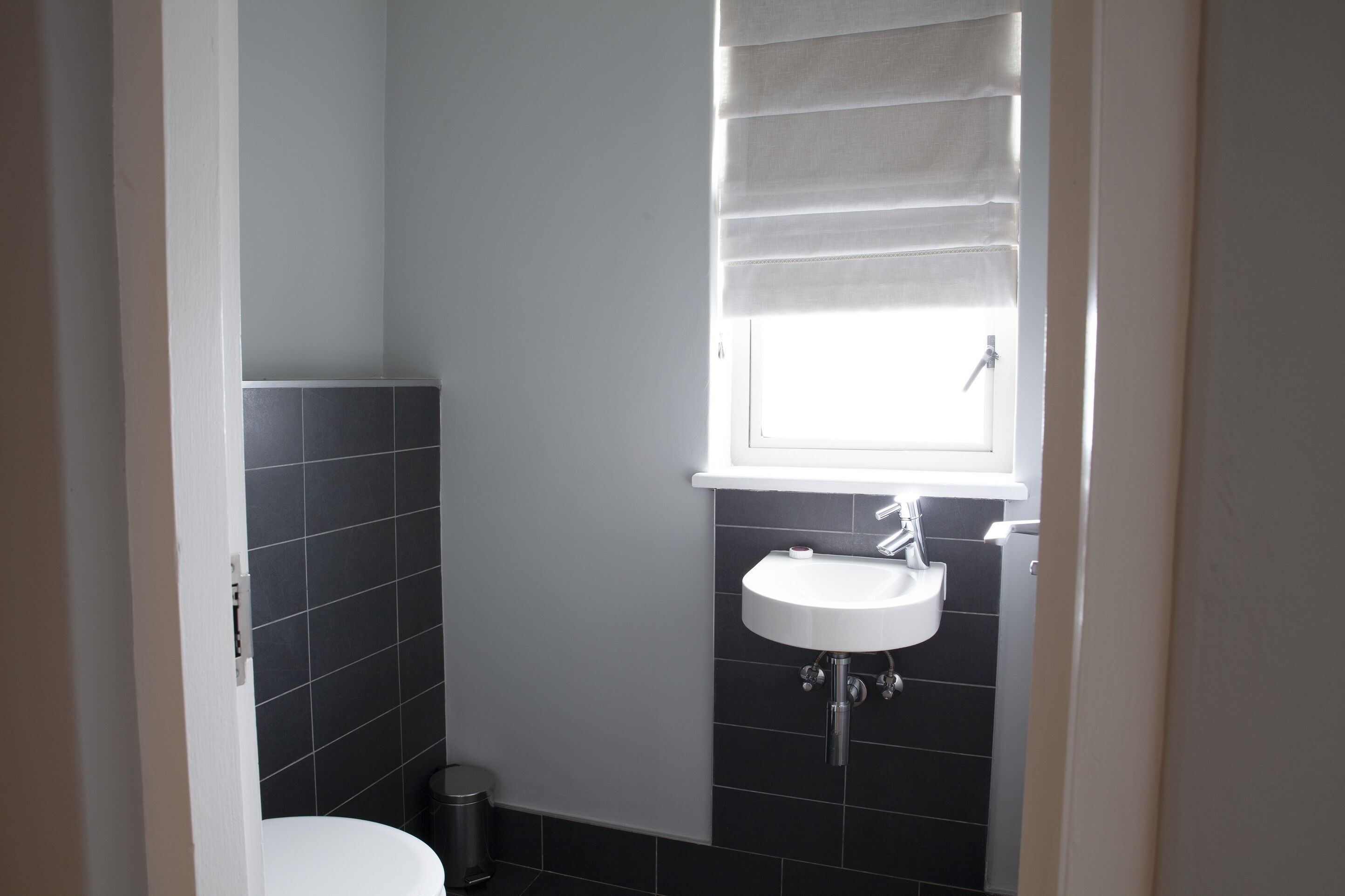 A bathroom with a white sink, a window with blinds, and dark tiled walls.