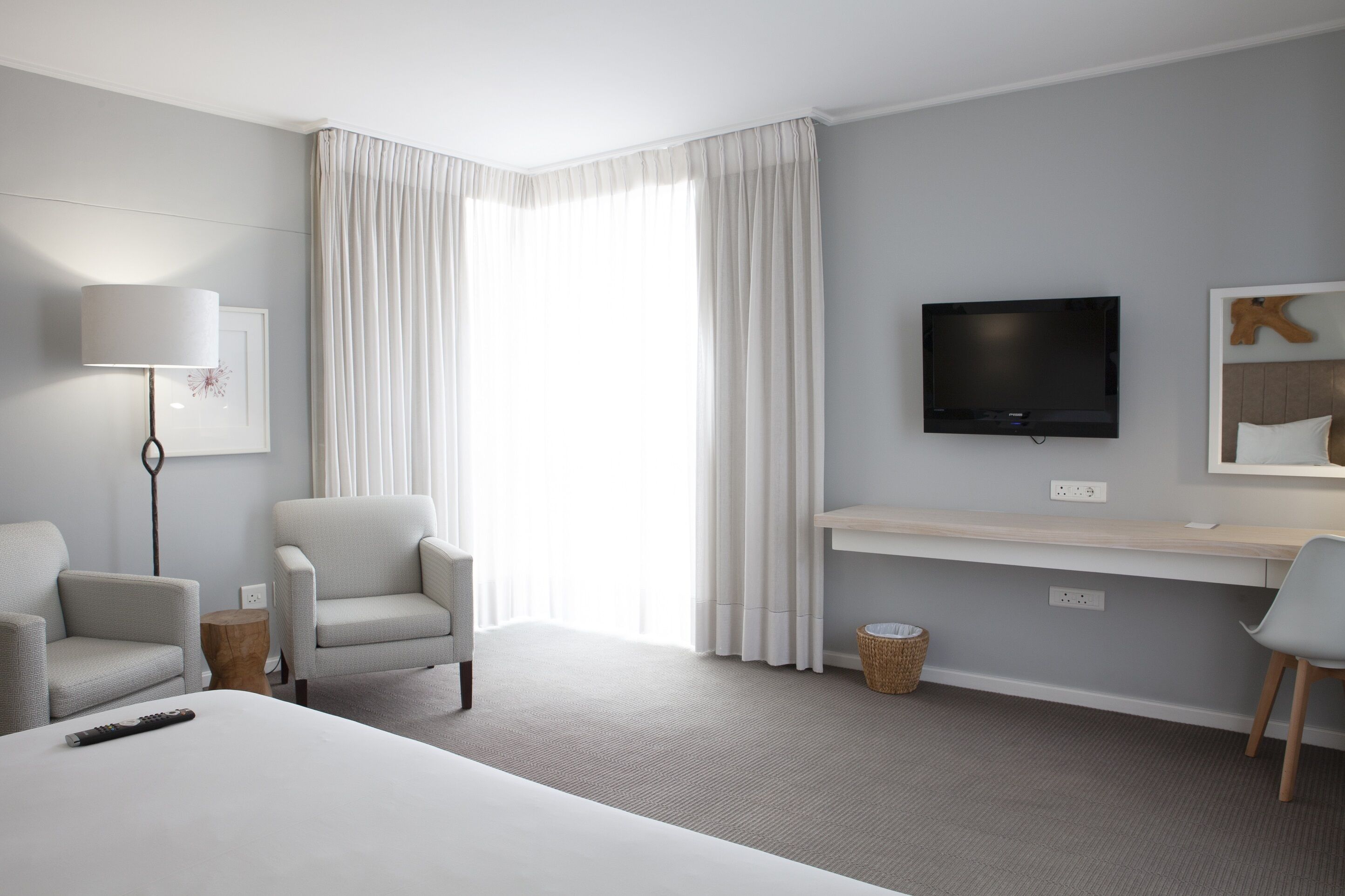 Quarto de hotel com cama, duas poltronas, escrivaninha com televisão e uma janela grande com cortinas.