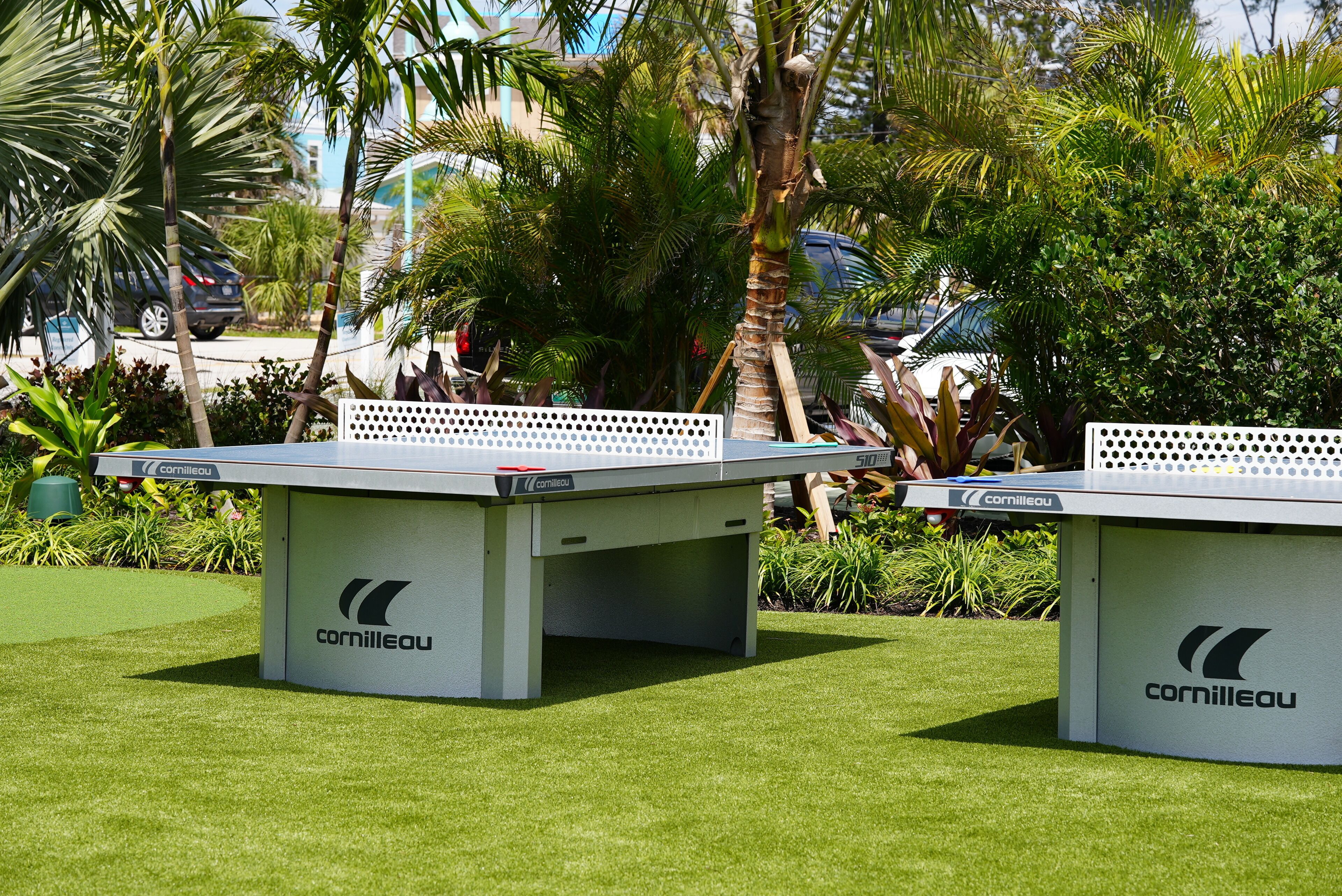 Two Cornilleau ping pong tables in an outdoor setting with greenery and parked cars in the background.