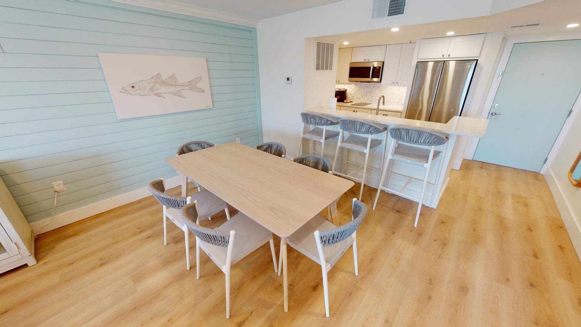 Une cuisine moderne dotée d’appareils électroménagers en acier inoxydable, un coin repas avec une table en bois et des chaises en osier, et un mur bleu clair orné d’une œuvre d’art représentant des poissons.