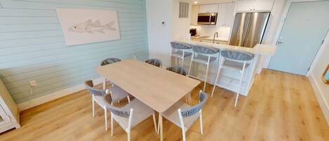 Une cuisine moderne dotée d’appareils électroménagers en acier inoxydable, un coin repas avec une table en bois et des chaises en osier, et un mur bleu clair orné d’une œuvre d’art représentant des poissons.