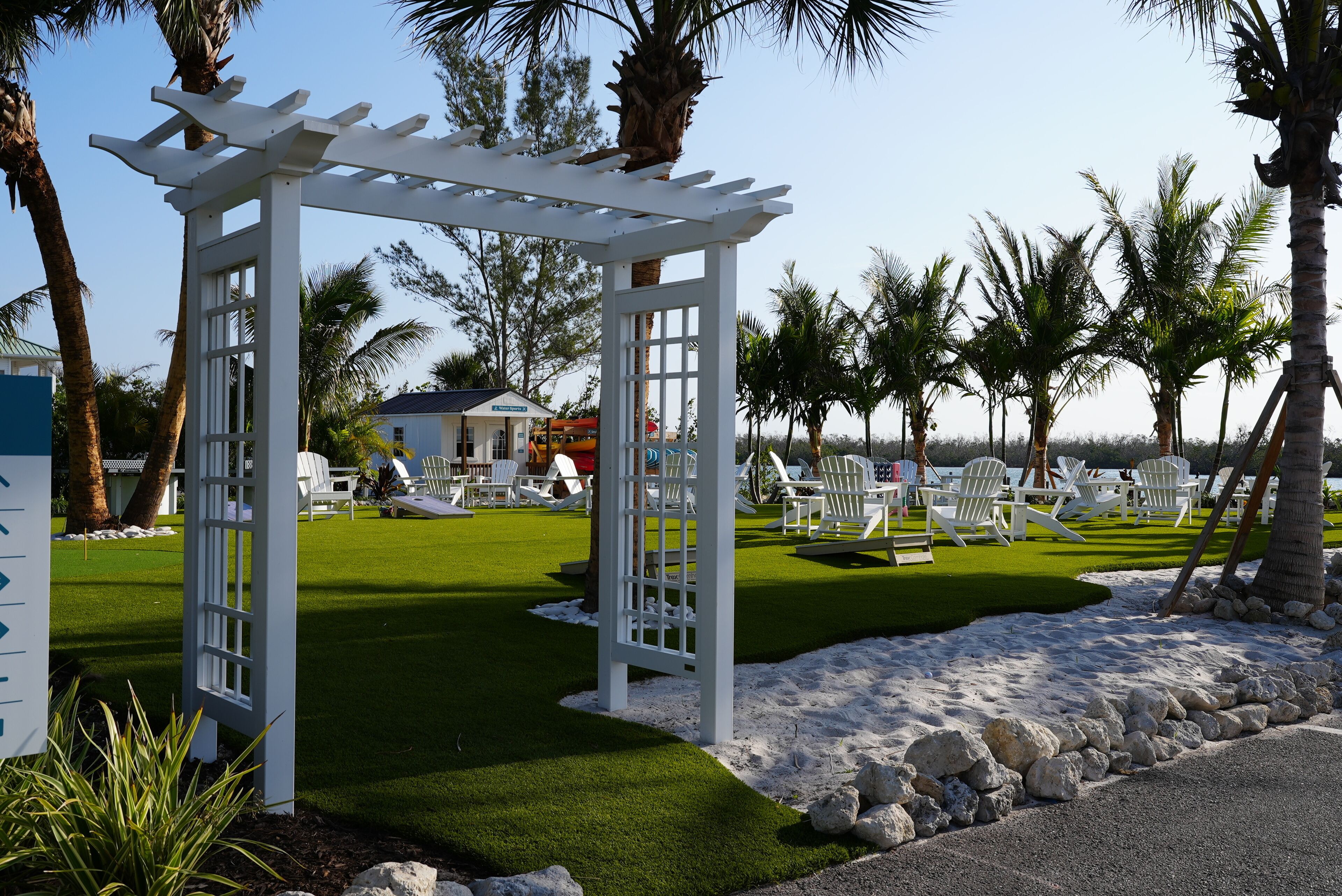 Eine weiße Pergola, helle Lounge-Stühle und ein gepflegter Rasen mit Palmen.