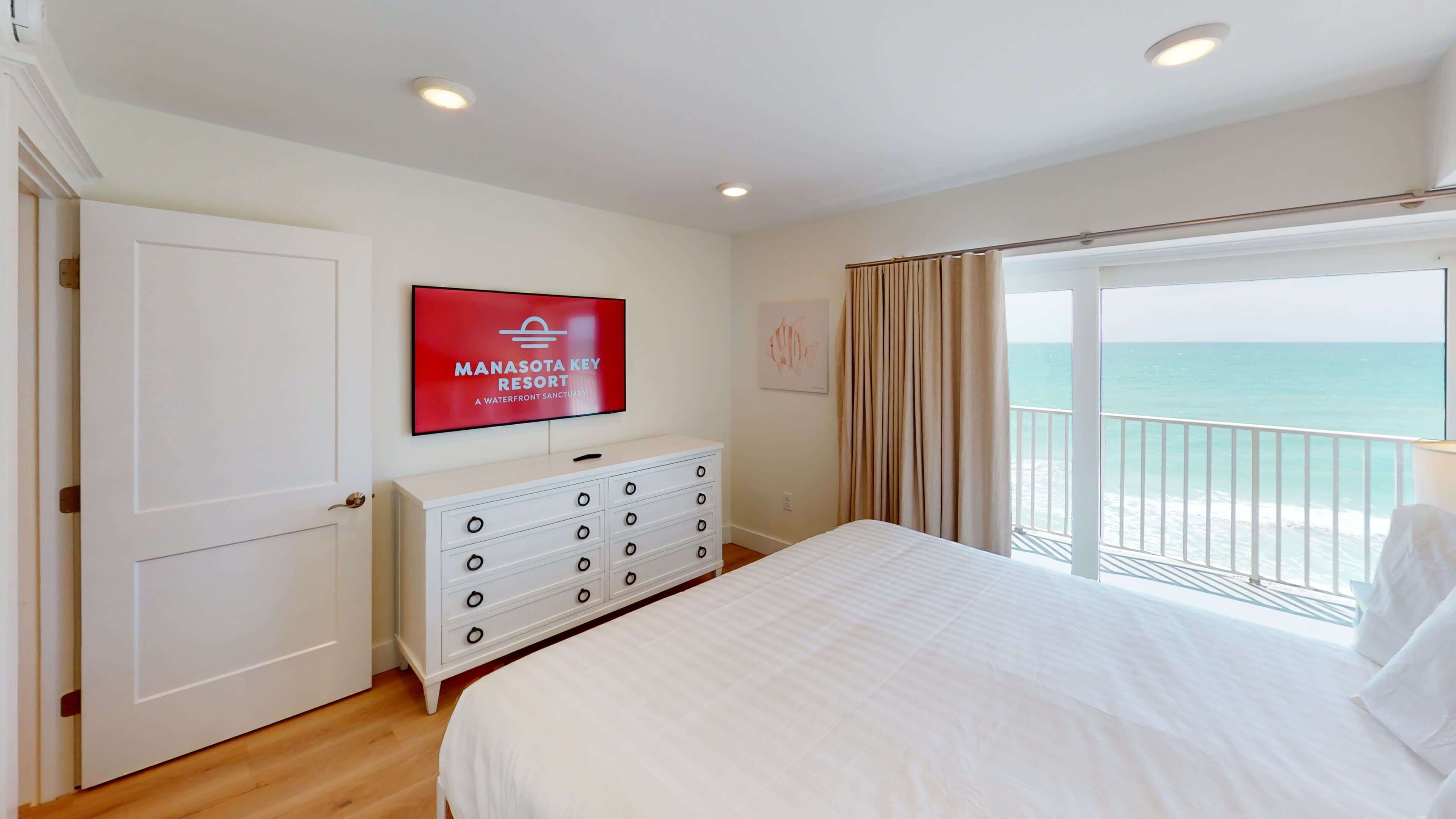 Une chambre d’hôtel avec un lit, une commode, un téléviseur diffusant des images du « Manasota Key Resort », une fenêtre avec des rideaux et une vue sur l’océan.