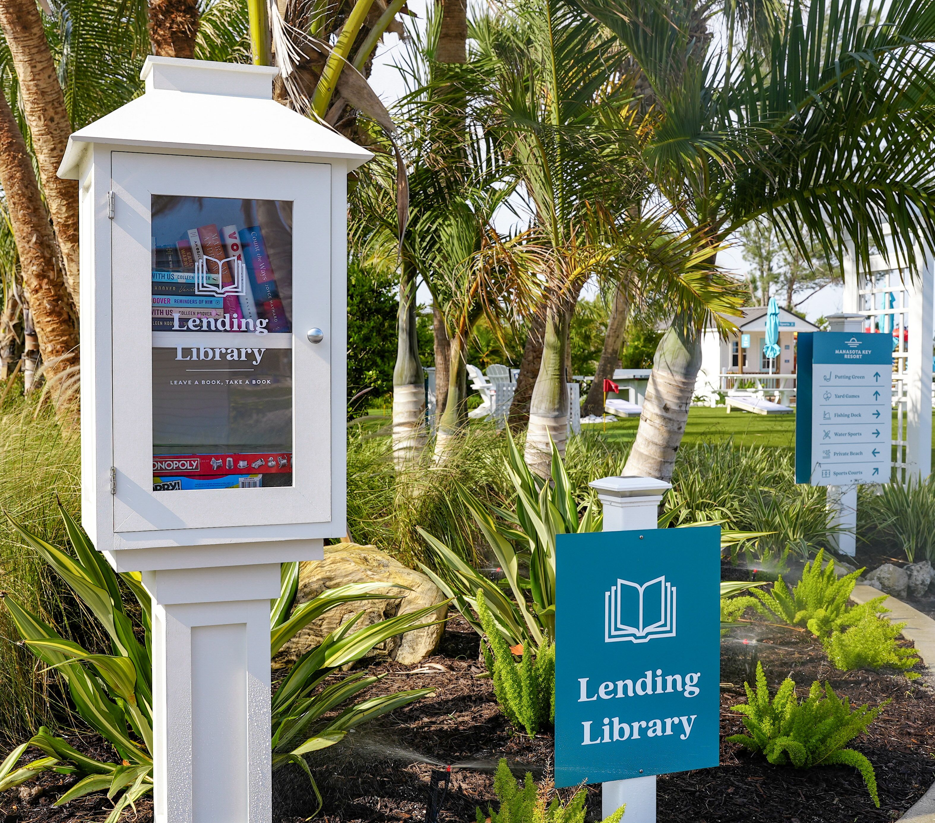 Schild für eine Leihbibliothek in einem Garten mit Palmen und einem weißen Gebäude.